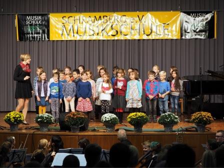 Ein KInderchor steht mit der Lehrerin auf der Bühne. Im Vordergrund das Orchester vor der Bühne.